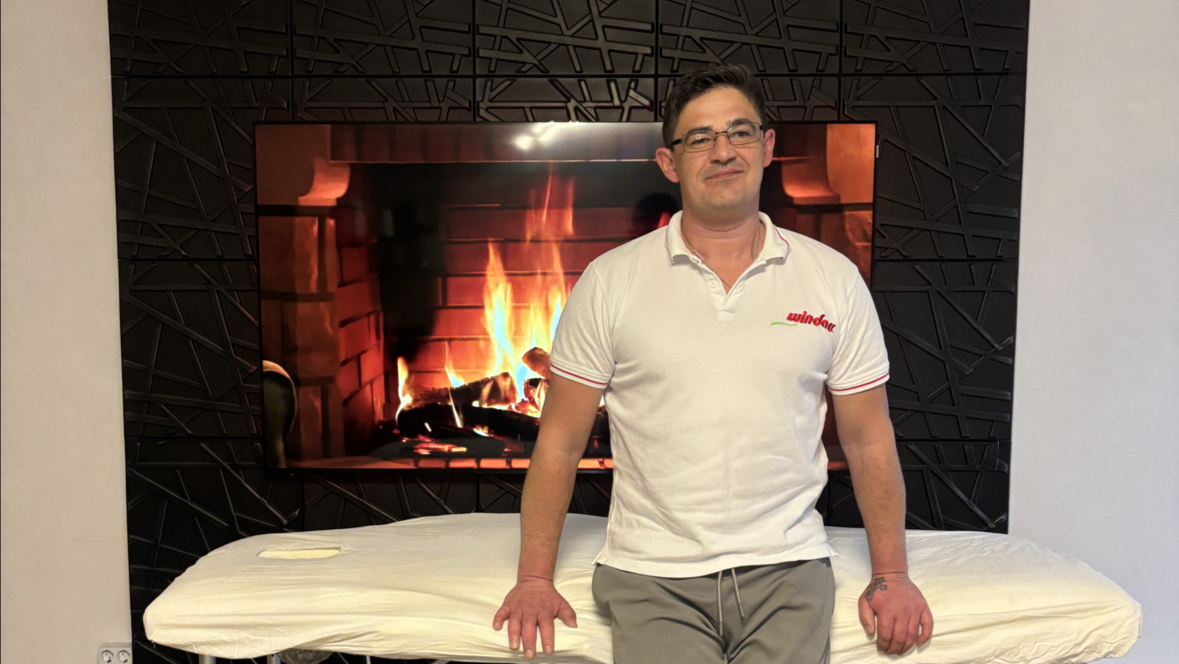 Denis Seibert bei seiner Massageliege vor dem Kaminmotiv